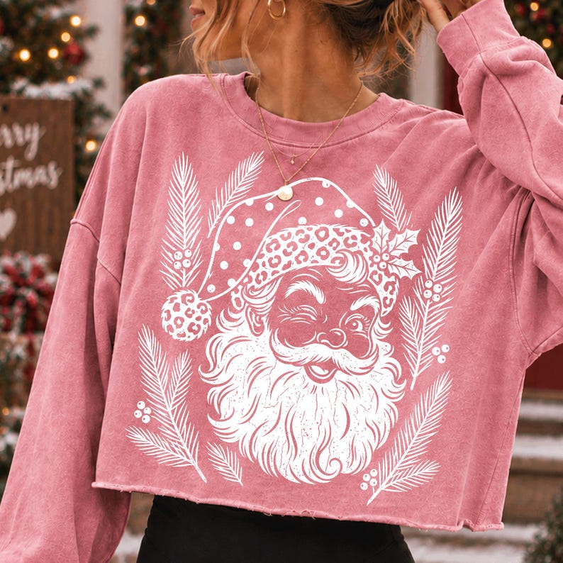 Puede incluir: Una sudadera corta de manga larga en color rosa empolvado con un estampado gr&aacute;fico blanco de un Santa Claus gui&ntilde;ando un ojo, con un gorro de lunares y estampado de leopardo, rodeado de ramas de pino y bayas de acebo estilizadas.