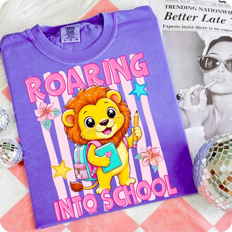 May include: A purple t-shirt featuring a cartoon lion holding a pencil and a book, with a pink backpack. The graphic includes the text "ROARING INTO SCHOOL" in pink letters, surrounded by pink and white vertical stripes, stars, and flowers.
