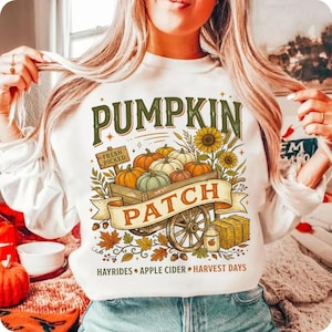 Puede incluir: Una sudadera blanca de manga larga con un gr&aacute;fico de un carro de madera lleno de calabazas, girasoles y una paca de heno. El texto dice PUMPKIN PATCH, FRESH PICKED, HAYRIDES, APPLE CIDER y HARVEST DAYS en colores verde y naranja.
