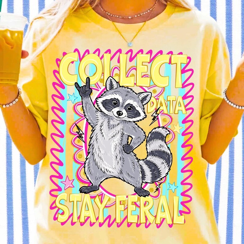 May include: Yellow t-shirt featuring a cartoon raccoon illustration. The raccoon is giving a hand gesture. The text on the shirt says "COLLECT DATA STAY FERAL" in a pink and yellow design.
