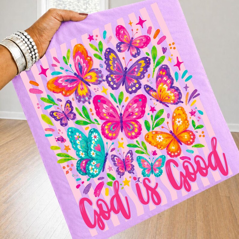 May include: A light purple fabric featuring a pattern of colorful butterflies in shades of pink, orange, yellow, and teal, accented with green leaves and small stars. The text "God is Good" is printed in a bold, cursive pink font at the bottom.