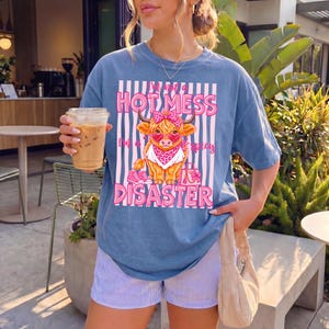 K&ouml;nnte beinhalten: Blaues T-Shirt mit einer Grafik einer Highland-Kuh mit rosa Schleife, Sonnenbrille und Bandana. Der Text lautet "I'm Not a Hot Mess, I'm a Spicy Disaster." Das Shirt wird mit gestreiften Shorts getragen.