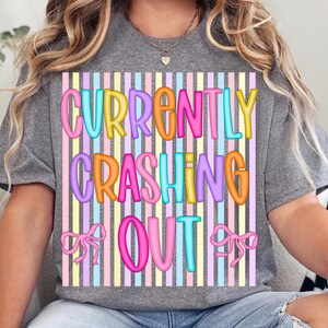 Pode incluir: Camiseta cinza com um gr&aacute;fico colorido que diz "CURRENTLY CRASHING OUT" em letras de bolhas. O design apresenta um fundo listrado em cores pastel e dois la&ccedil;os rosa.