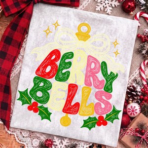 May include: A light gray t-shirt featuring a graphic of a Christmas ornament shaped like a bell with the text BERRY BELLS. The design includes red, green, and yellow scribbled lettering, a yellow bow, and green holly leaves with red berries.