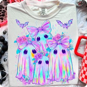 May include: Cream-colored t-shirt featuring a whimsical design of three pastel-colored ghosts with large bows and floral accents. Two purple bats are above the ghosts. The shirt has a Comfort Colors tag.