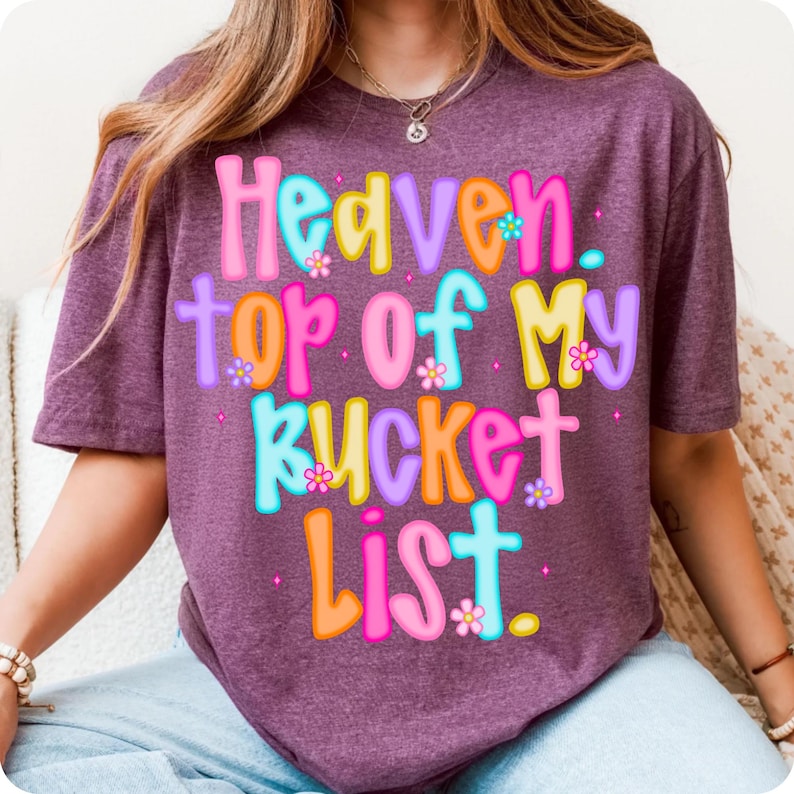 K&ouml;nnte beinhalten: Ein weinrotes T-Shirt mit dem Text "Heaven, top of my bucket list" in bunten Buchstaben. Der Text ist mit kleinen Blumen verziert. Es ist ein l&auml;ssiges Kleidungsst&uuml;ck.