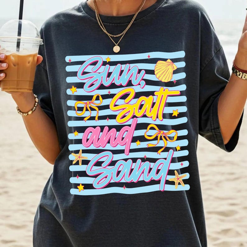 Peut inclure: T-shirt gris fonc&eacute; avec l'inscription "Sun, Salt, and Sand" dans un design color&eacute;. Le motif comprend des rayures bleues, des &eacute;toiles jaunes, un coquillage, des n&oelig;uds et des &eacute;toiles de mer. V&ecirc;tement d&eacute;contract&eacute; sur le th&egrave;me de la plage.