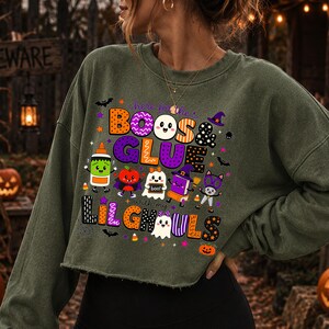 K&ouml;nnte beinhalten: Ein olivgr&uuml;nes, lang&auml;rmeliges, bauchfreies Sweatshirt mit einer bunten Halloween-Grafik und dem Text "here for the BOOS & GLUE with my LIL GHOULS". Das Design zeigt Zeichentrickfiguren wie Geister, eine Klebeflasche und ein Buch.