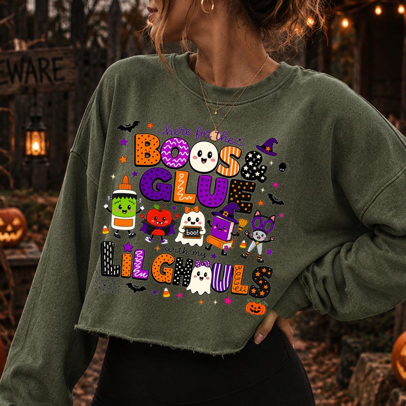 K&ouml;nnte beinhalten: Ein olivgr&uuml;nes, lang&auml;rmeliges, bauchfreies Sweatshirt mit einer bunten Halloween-Grafik und dem Text "here for the BOOS & GLUE with my LIL GHOULS". Das Design zeigt Zeichentrickfiguren wie Geister, eine Klebeflasche und ein Buch.