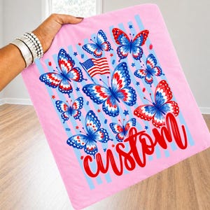 Peut inclure: Un carr&eacute; de tissu rose clair orn&eacute; de papillons aux ailes rouges, blanches et bleues. Le motif comprend un petit drapeau am&eacute;ricain, des &eacute;toiles bleues dispers&eacute;es et des rayures verticales bleu clair. Le mot custom est &eacute;crit en rouge.