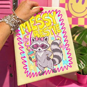 以下が含まれることがあります： ハート型のサングラスとピンクのリボンをつけた漫画のアライグマが描かれた、明るい黄色のトートバッグ。「Messy Bestie」の文字は黄色でピンクの縁取り。アライグマはハートのマークが入った飲み物を持っています。