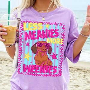 May include: A lilac t-shirt with a graphic of a dachshund wearing sunglasses. The text "LESS MEANIES MORE WEENIES" is in yellow and pink, with stars and a blue striped background. A fun, casual design.
