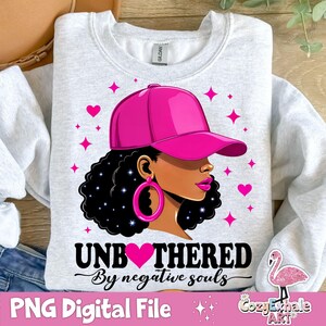 May include: A white sweatshirt featuring a pink graphic of a woman wearing a pink baseball cap and large hoop earrings. The graphic includes the text "Unbothered by negative souls".