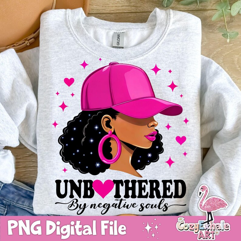May include: A white sweatshirt featuring a pink graphic of a woman wearing a pink baseball cap and large hoop earrings. The graphic includes the text "Unbothered by negative souls".