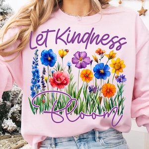 May include: A light pink sweatshirt with the words "Let Kindness Bloom" in purple script. The design features a colorful floral arrangement with blue, yellow, red, and orange flowers, and green grass.