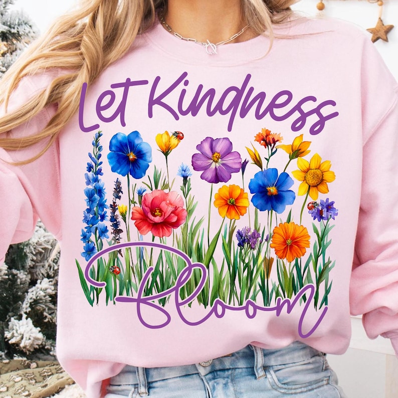 May include: A light pink sweatshirt with the words "Let Kindness Bloom" in purple script. The design features a colorful floral arrangement with blue, yellow, red, and orange flowers, and green grass.