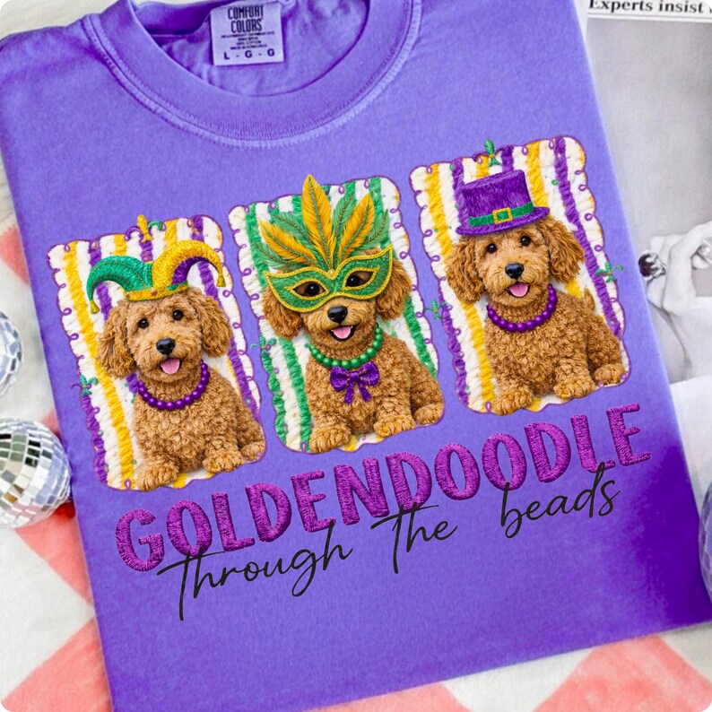 May include: A purple t-shirt featuring three Goldendoodle puppies wearing Mardi Gras attire. Each dog has a different hat or mask and a beaded necklace. The text "GOLDENDOODLE Through The beads" is below the dogs.