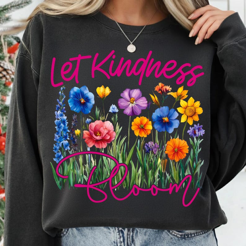May include: Dark gray sweatshirt with the words "Let Kindness Bloom" in pink script. The design features a colorful floral arrangement with blue, pink, yellow, orange, and purple flowers, and green grass.
