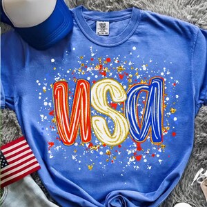 Peut inclure: Un t-shirt bleu avec le mot "USA" en rouge, blanc et bleu, avec un motif &eacute;clabouss&eacute;. Un drapeau am&eacute;ricain et une casquette de baseball bleue et blanche sont &eacute;galement visibles.