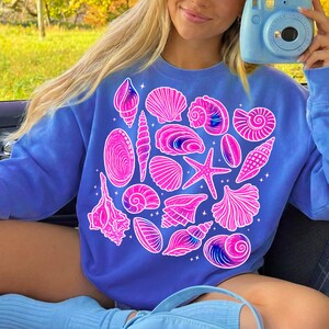 Pu&ograve; includere: Una felpa blu con un design di conchiglie rosa. Il disegno include varie forme di conchiglie e una stella marina, evocando un tema costiero. La felpa &egrave; di colore blu unito.
