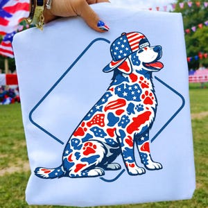 May include: A light blue t-shirt featuring a patriotic dog design. The dog is wearing a baseball cap and is decorated with red, white, and blue stars and stripes. The design is set within a blue diamond shape.