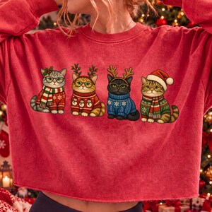 K&ouml;nnte beinhalten: Ein rotes Langarm-Sweatshirt mit einem Grafikdruck von vier Katzen, die weihnachtliche Pullover, Schals und Accessoires tragen. Die Katzen sind mit Brillen, Rentiergeweihen und einer Weihnachtsm&uuml;tze in einer horizontalen Reihe abgebildet.