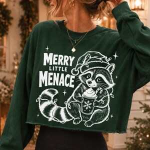 May include: A dark green long-sleeve cropped sweatshirt featuring a white graphic of a raccoon wearing a Santa hat and holding a cup with a candy cane. The text reads Merry Little Menace, surrounded by small star accents and string lights.
