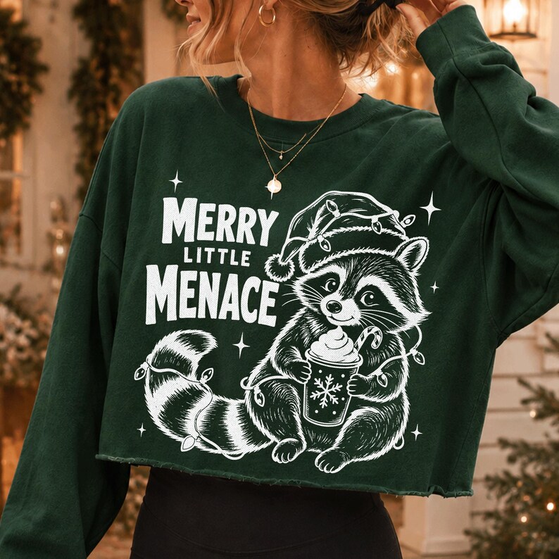 May include: A dark green long-sleeve cropped sweatshirt featuring a white graphic of a raccoon wearing a Santa hat and holding a cup with a candy cane. The text reads Merry Little Menace, surrounded by small star accents and string lights.