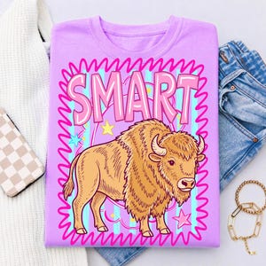 May include: A lilac-colored t-shirt featuring a cartoon bison illustration. The word "SMART" is displayed in pink bubble letters within a pink wavy border. The bison is tan with a detailed mane and horns. Stars and stripes are in the background.