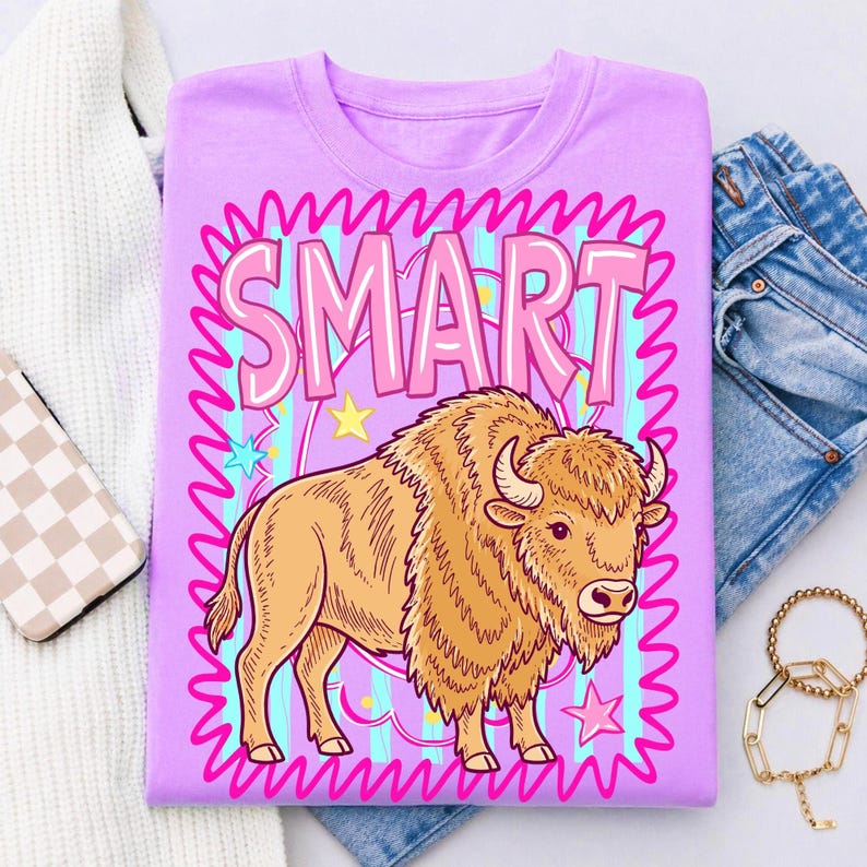 May include: A lilac-colored t-shirt featuring a cartoon bison illustration. The word "SMART" is displayed in pink bubble letters within a pink wavy border. The bison is tan with a detailed mane and horns. Stars and stripes are in the background.