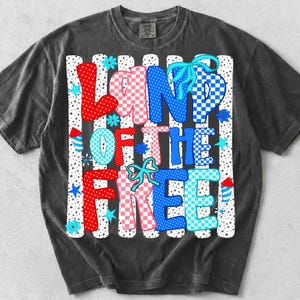 Puede incluir: Una camiseta gris carb&oacute;n con un estampado gr&aacute;fico que muestra el texto LAND OF THE FREE en patrones de cuadros y lunares rojos, blancos y azules. El dise&ntilde;o incluye lazos azul claro, flores peque&ntilde;as, estrellas e ilustraciones de fuegos artificiales.