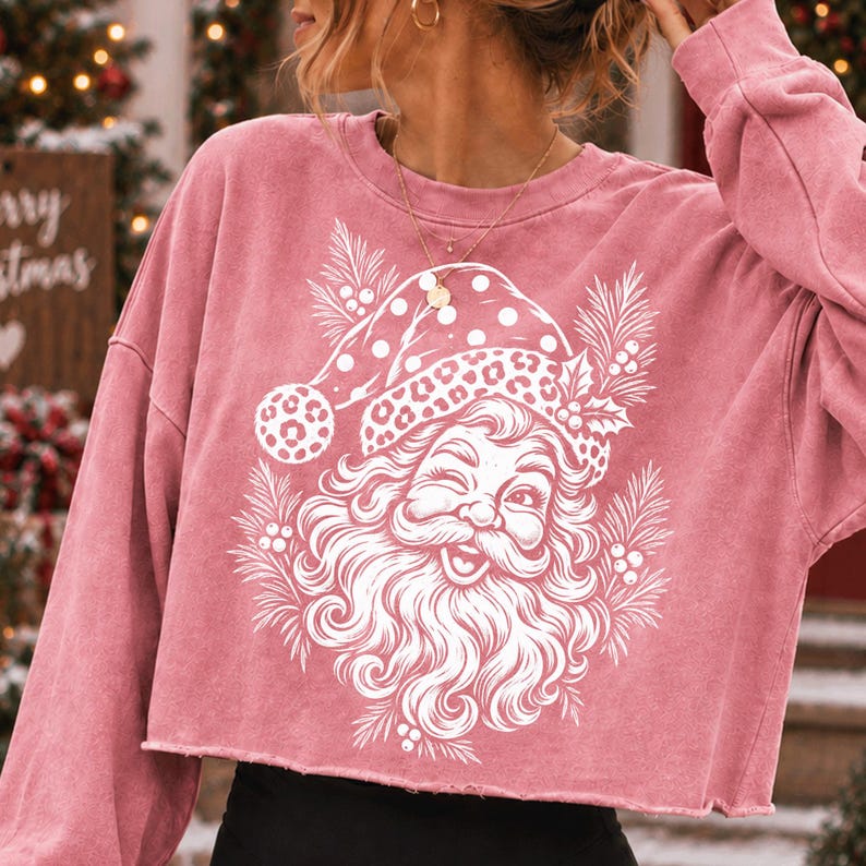 K&ouml;nnte beinhalten: Ein altrosa lang&auml;rmeliges bauchfreies Sweatshirt mit einem wei&szlig;en Grafikdruck eines zwinkernden Weihnachtsmanns, der eine M&uuml;tze mit Leopardenmuster tr&auml;gt, umgeben von Tannennadeln und Stechpalmenbeeren, mit einem rohen, unges&auml;umten Saum.