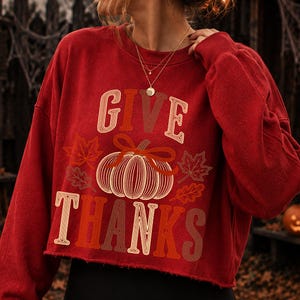 Puede incluir: Una sudadera roja de manga larga y corte corto con un gr&aacute;fico de una calabaza con un lazo y hojas de oto&ntilde;o. El texto "GIVE THANKS" est&aacute; impreso en una fuente retro multicolor sobre y debajo de la ilustraci&oacute;n de la calabaza en tonos crema y marr&oacute;n.