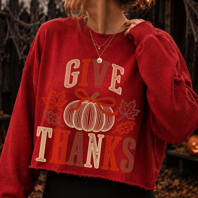 Puede incluir: Una sudadera roja de manga larga y corte corto con un gr&aacute;fico de una calabaza con un lazo y hojas de oto&ntilde;o. El texto "GIVE THANKS" est&aacute; impreso en una fuente retro multicolor sobre y debajo de la ilustraci&oacute;n de la calabaza en tonos crema y marr&oacute;n.