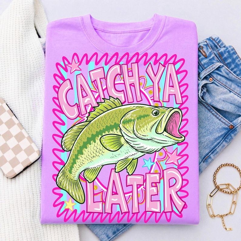 Op de afbeelding: Een lichtpaars T-shirt met een groen en gele visafbeelding en de tekst "CATCH YA LATER" in roze en blauw. Het ontwerp bevat sterren en een golvende roze rand. Het shirt wordt getoond met een spijkerbroek en een witte trui.