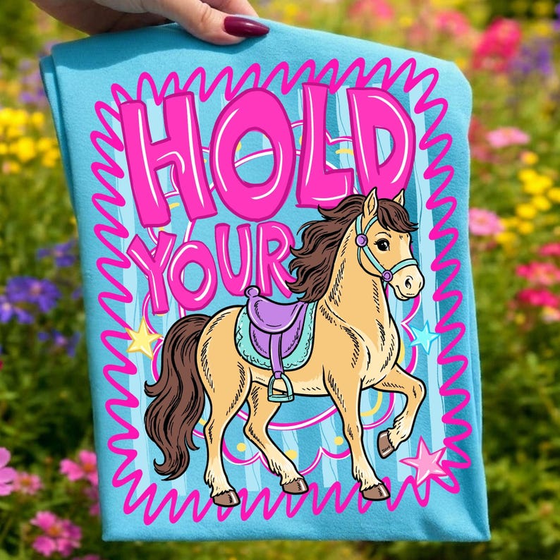 May include: A light blue t-shirt featuring a cartoon horse with a purple saddle and the text "HOLD YOUR". The design includes pink stars and a pink wavy border. The horse is tan with a brown mane and tail.