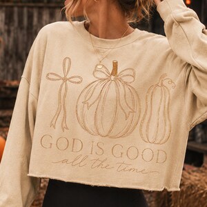 K&ouml;nnte beinhalten: Ein beigefarbenes, lang&auml;rmeliges bauchfreies Sweatshirt mit einer braunen Strichzeichnung einer Schleife, eines K&uuml;rbisses mit Schleife und einer K&uuml;rbisfrucht. Der Text "GOD IS GOOD" steht in serifenbetonter Schrift, darunter "all the time".