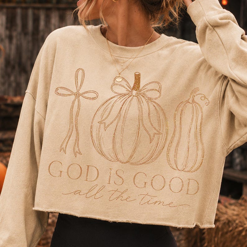 K&ouml;nnte beinhalten: Ein beigefarbenes, lang&auml;rmeliges bauchfreies Sweatshirt mit einer braunen Strichzeichnung einer Schleife, eines K&uuml;rbisses mit Schleife und einer K&uuml;rbisfrucht. Der Text "GOD IS GOOD" steht in serifenbetonter Schrift, darunter "all the time".