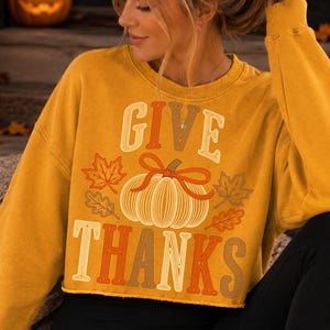 Puede incluir: Una sudadera de manga larga color amarillo mostaza con un estampado gr&aacute;fico de una calabaza con un lazo encima, rodeada de hojas de oto&ntilde;o. El texto "GIVE" est&aacute; impreso sobre la calabaza y "THANKS" debajo en una fuente estilo retro.