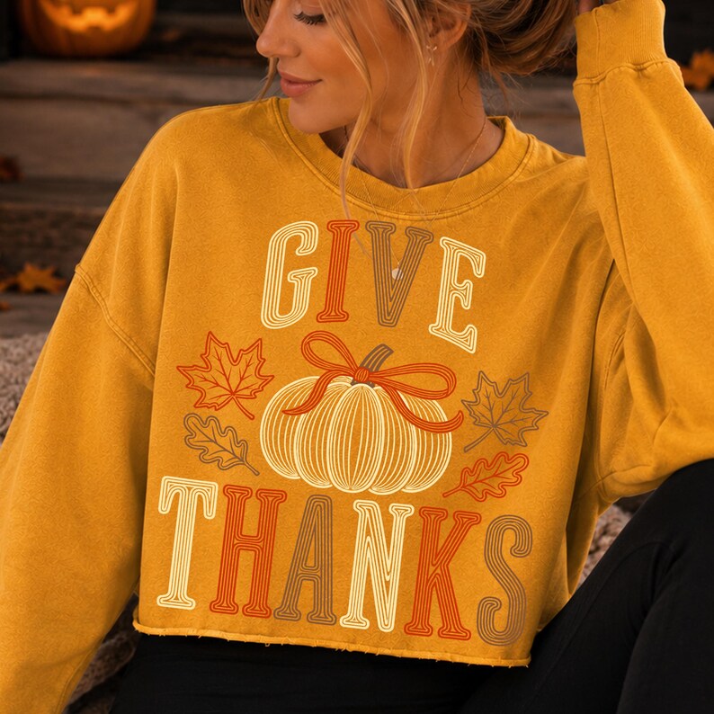 Puede incluir: Una sudadera de manga larga color amarillo mostaza con un estampado gr&aacute;fico de una calabaza con un lazo encima, rodeada de hojas de oto&ntilde;o. El texto "GIVE" est&aacute; impreso sobre la calabaza y "THANKS" debajo en una fuente estilo retro.