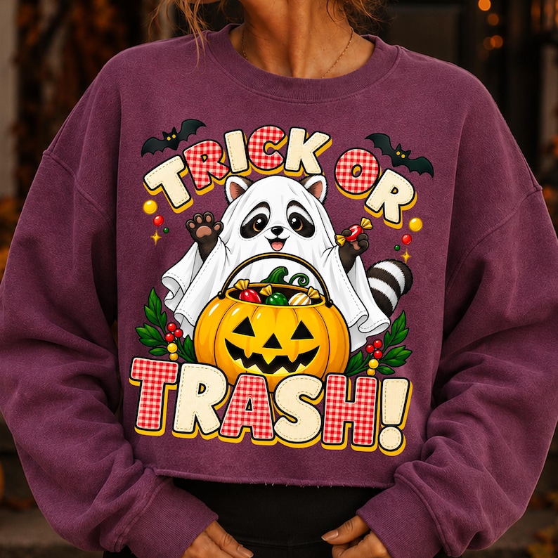 May include: A dark purple long-sleeve sweatshirt featuring a graphic of a raccoon wearing a white ghost costume, holding a jack-o'-lantern bucket filled with sweets. The text reads TRICK OR TRASH! with bats and red and white checkered accents.