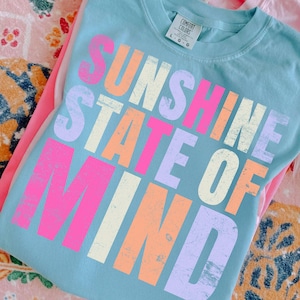 May include: A light blue Comfort Colors sweatshirt with the words "SUNSHINE STATE OF MIND" in a colourful, distressed font. The text is in shades of pink, orange, purple, and cream. The sweatshirt is folded on top of a pink shirt.