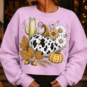 Puede incluir: Una sudadera de manga larga color morado claro con un estampado gr&aacute;fico de una calabaza con manchas de vaca, una calabaza de cuadros amarillos, un cactus verde con flores blancas, hojas de oto&ntilde;o, bellotas, trigo y margaritas blancas.