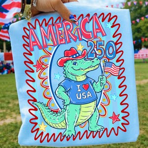 Puede incluir: Camiseta azul claro con un cocodrilo de dibujos animados que lleva un sombrero de vaquero y una camiseta azul con "I ❤️ USA". La camiseta tiene el texto "AMERICA 250" en rojo y azul, con un borde decorativo. El cocodrilo sostiene una bandera americana.