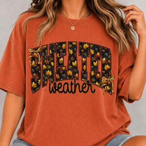 May include: A burnt orange short-sleeve t-shirt featuring the text "SWEATER weather" in a bold, blocky font filled with a pattern of yellow and orange autumn leaves. The design includes two small yellow bows and tiny star accents.