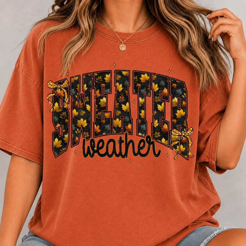 May include: A burnt orange short-sleeve t-shirt featuring the text "SWEATER weather" in a bold, blocky font filled with a pattern of yellow and orange autumn leaves. The design includes two small yellow bows and tiny star accents.