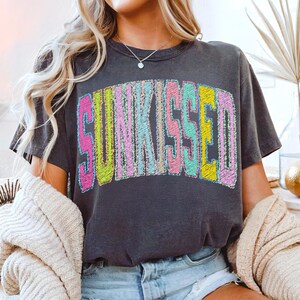May include: Dark grey t-shirt with the word "SUNKISSED" in colourful, embroidered-style lettering. The text features pink, yellow, blue, and green hues. The shirt has a crew neck and short sleeves.