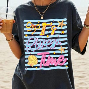 May include: Dark gray t-shirt with a graphic that says "It's River Time" in yellow, blue, and pink. The design includes a bow, starfish, shell, and stars on blue and white stripes. The shirt is a casual, beach-themed design.