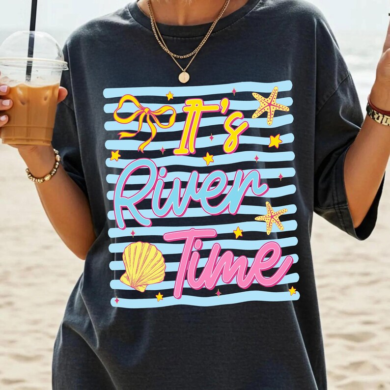 May include: Dark gray t-shirt with a graphic that says "It's River Time" in yellow, blue, and pink. The design includes a bow, starfish, shell, and stars on blue and white stripes. The shirt is a casual, beach-themed design.