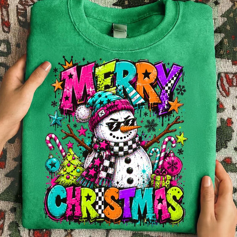 Puede incluir: Una sudadera verde brillante de cuello redondo con un gr&aacute;fico de un mu&ntilde;eco de nieve con gafas de sol, un gorro turquesa y rosa, y una bufanda a cuadros. El texto MERRY CHRISTMAS est&aacute; impreso en letras coloridas estilo grafiti con estrellas.